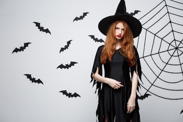 Halloween witch concept  halloween witch holding posing with serious expression over dark grey  wall with bat and spider web