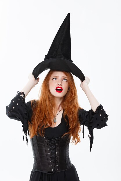 Концепция ведьмы Хэллоуина - Счастливый Хэллоуин Ведьма с сексуальными рыжими волосами борется со своей волшебной шляпой, пролетая над ее головой. Изолированный на белой стене.
