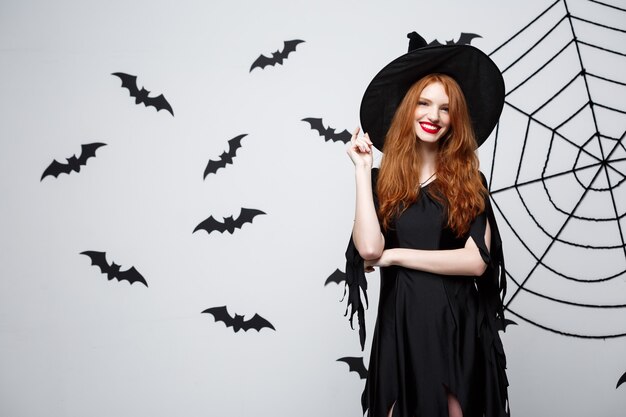 Halloween witch concept  happy halloween witch holding posing over dark grey  wall with bat and spider web