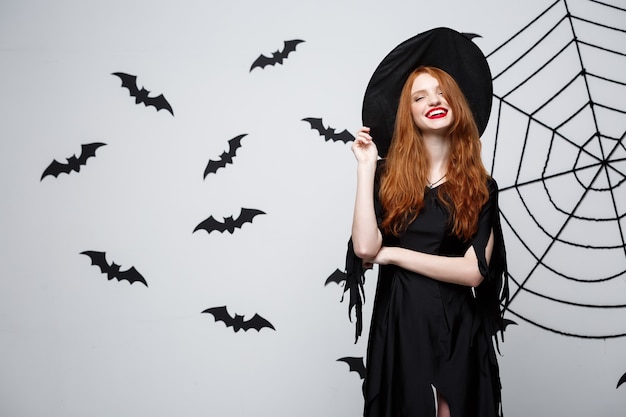 Бесплатное фото Хэллоуин ведьма концепция счастливая хэллоуин ведьма держит позирует над темно-серой стеной с летучей мышью и паутиной
