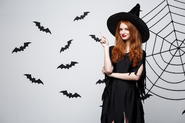 Halloween witch concept  happy halloween witch pointing finger on side over dark grey  wall with bat and spider web