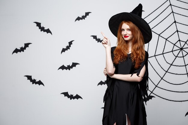 Halloween witch concept  happy halloween witch pointing finger on side over dark grey  wall with bat and spider web