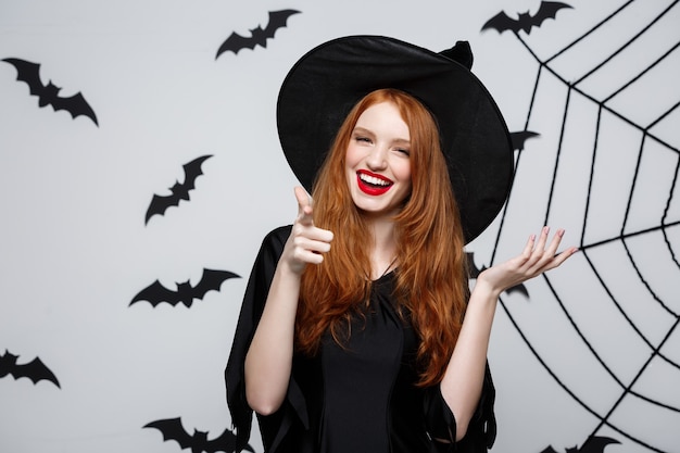 Halloween witch concept - Happy Halloween Witch pointing finger on side over dark grey  wall with bat and spider web.