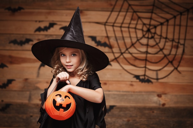 Halloween witch concept - little caucasian witch child enjoy with halloween candy pumpkin jar. over bat and spider web background.