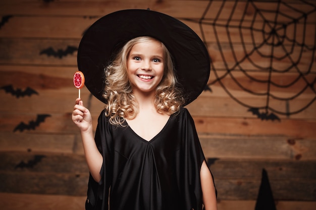 ハロウィーンの魔女のコンセプト-陽気な笑顔でハロウィーンの甘いとキャンディーを持つ小さな魔女の子供。バットとクモの巣の背景の上。