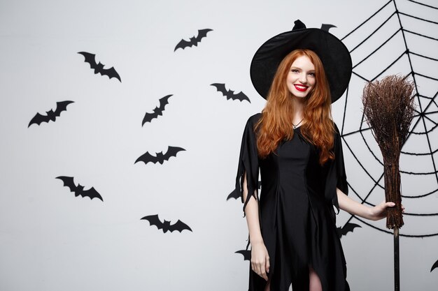 Halloween witch concept  portrait of beautiful young witch with broomstick over grey wall with bat and spider web wall