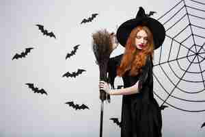 Free photo halloween witch concept  portrait of beautiful young witch with broomstick over grey wall with bat and spider web wall