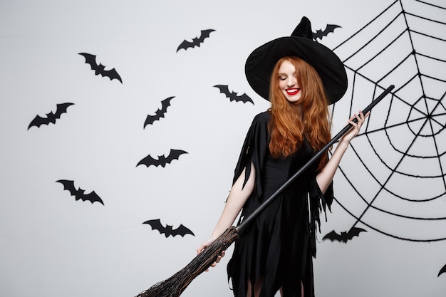 Halloween witch concept  portrait of beautiful young witch with broomstick over grey wall with bat and spider web wall