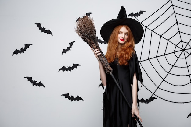 Бесплатное фото Хэллоуин ведьмы концептуальный портрет красивой молодой ведьмы с метлой над серой стеной с летучей мышью и стеной паутины