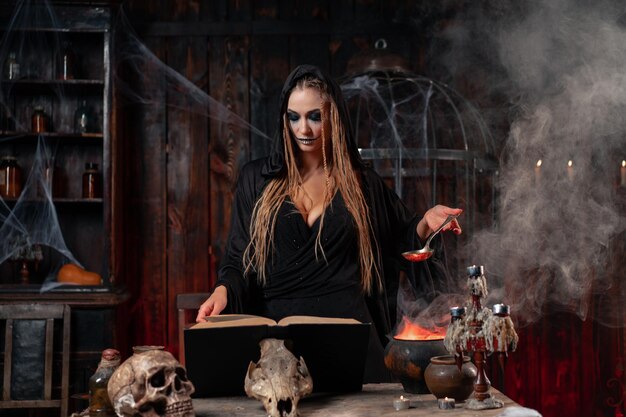 Halloween, witch reading magic book prepare poison or love potion in cauldron. black magic occult female wizard in dark room cage spider web human skull. necromancer cooking magic potion. voodoo magic