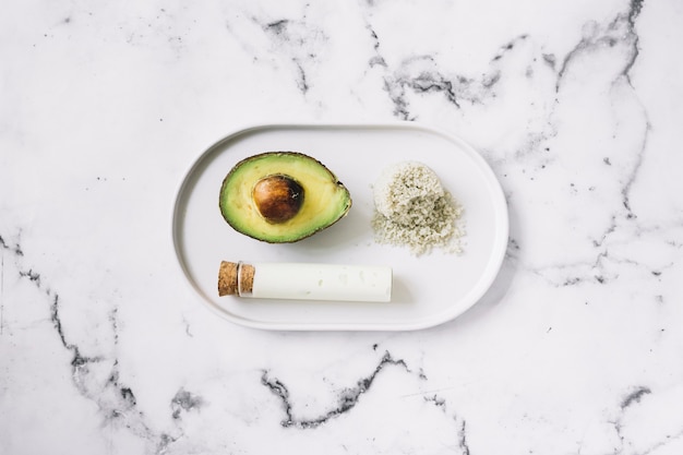 Halved avocado; grated body scrub and test tube on white tray against marble textured background