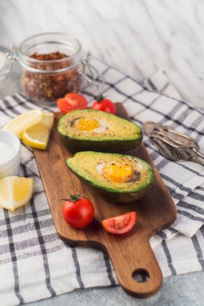 Halved avocado with egg york; tomatoes; lemon slice on chopping board over the napkin