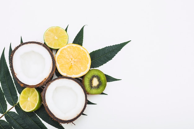 Halved coconut; lemon; orange and kiwi on green leaves against white background