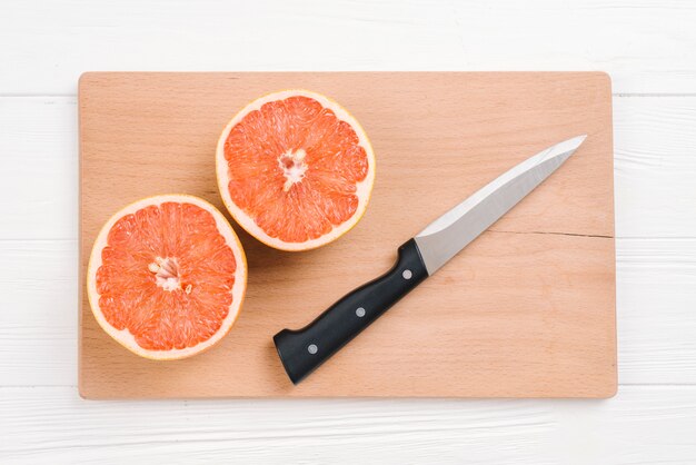 Halved grapefruits with sharp knife on wooden chopping board over white desk