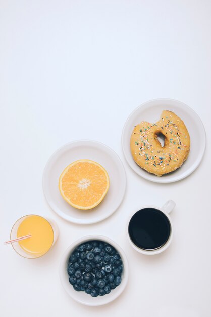 Половинный апельсин; апельсиновый сок; черника; чашка кофе и пончик на белом фоне