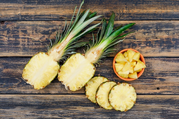 Halves and slices of pineapple in a orange bowl on old wood grunge, top view.