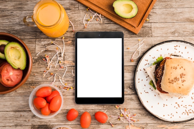 Hamburger; juice jar; tomatoes; avocado and sprout and digital tablet on wooden table