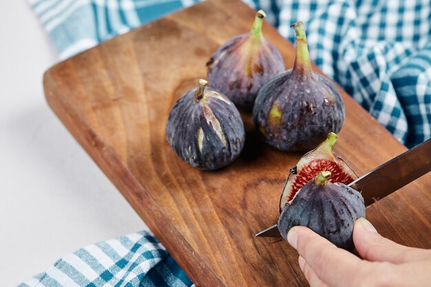 Hand cutting a fig on wooden cutting board with a blue tablecloth.