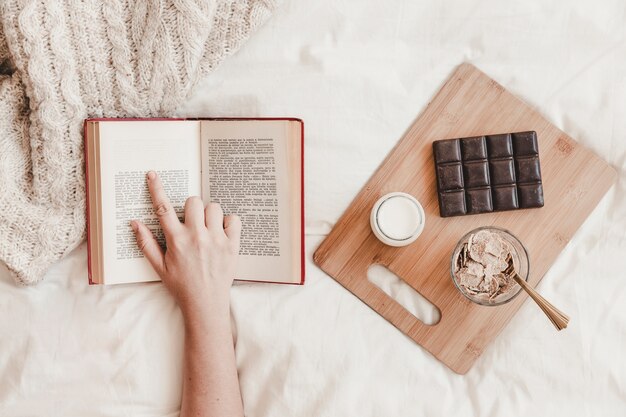 Hand following text in book near meal on bed