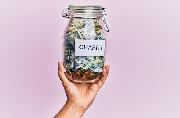 Free photo hand of hispanic man holding charity jar with dollars over isolated pink background