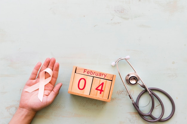 Hand holding awareness ribbon with stethoscope and 4th February wooden box on backdrop