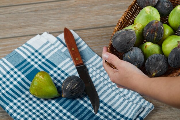 Hand holding a basket of figs on wooden table with a knife and tablecloth.