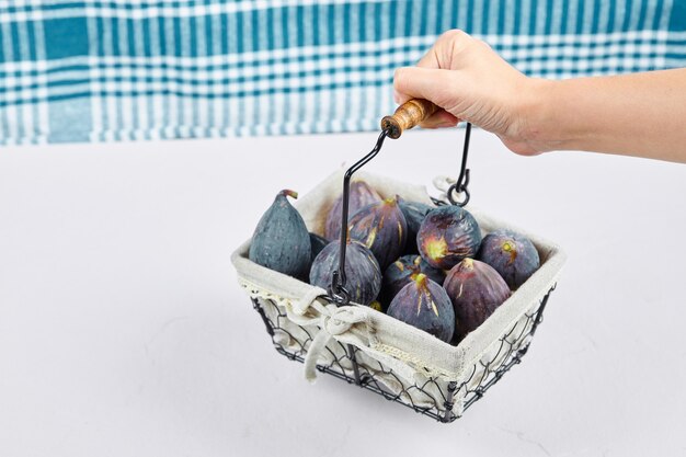 Hand holding a basket of purple figs on white with a blue tablecloth.