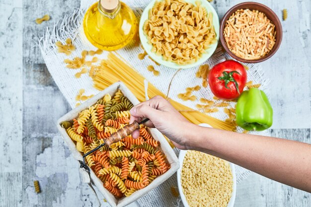 Hand holding a basket of raw fusilli pasta with assorted pastas and vegetable on the marble table.