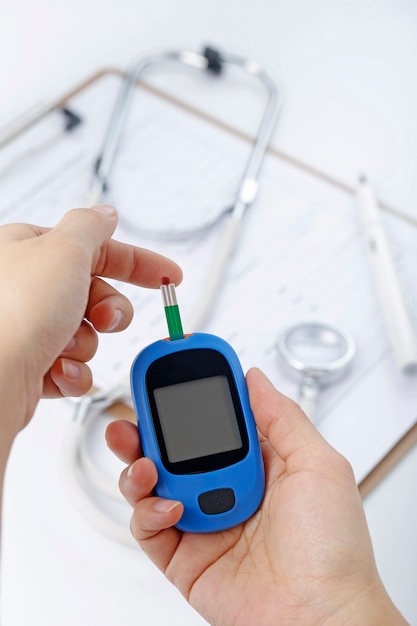 Hand holding a blood glucose meter measuring blood sugar, the background is a stethoscope and chart file