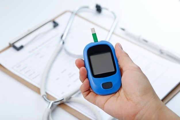Hand holding a blood glucose meter measuring blood sugar, the background is a stethoscope and chart file