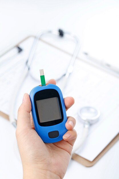 Hand holding a blood glucose meter measuring blood sugar, the background is a stethoscope and chart file