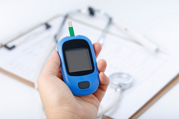 Hand holding a blood glucose meter measuring blood sugar, the background is a stethoscope and chart file