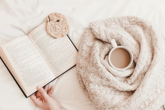 Hand holding book with biscuit on it near warm drink in plaid