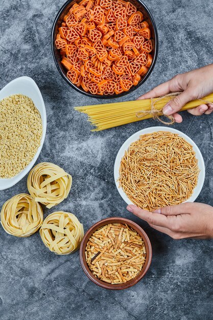 Hand holding a bowl of dry pasta with various types of raw pasta on the marble table.