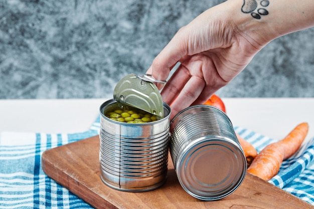 Hand holding a can of boiled green peas on a white table with vegetables and tablecloth.