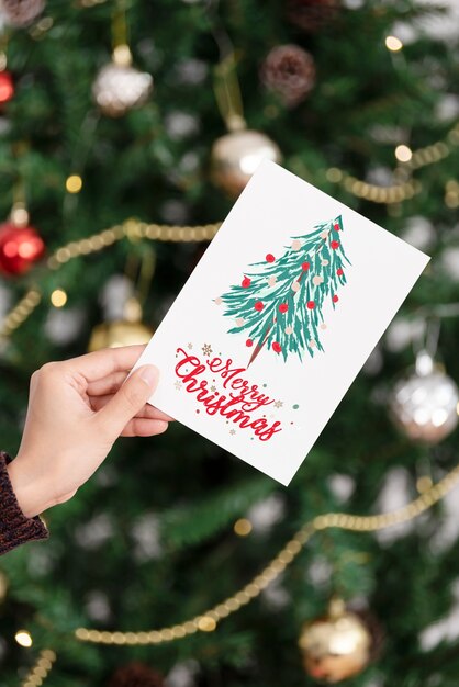 크리스마스 트리 배경에 크리스마스 인사말 카드를 들고 있는 손.