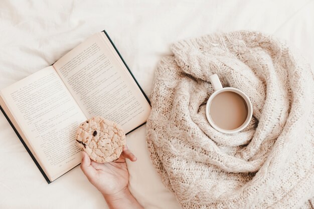 Hand holding cookie on book near hot drink in plaid