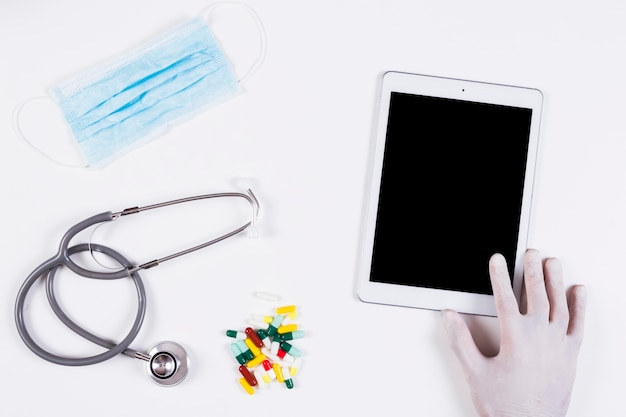 Hand holding digital tablet with mask; stethoscope and colorful capsules on white background