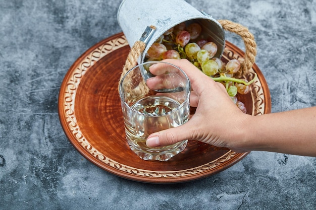 Free photo hand holding a glass of white wine and small bucket of grapes on a marble background. high quality photo