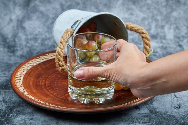 Hand holding a glass of white wine and small bucket of grapes on a marble surface.