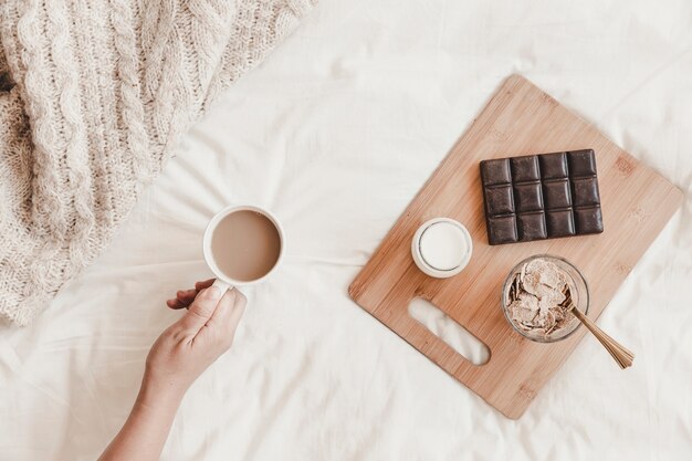 Hand holding hot drink near tasty meal on bedsheet