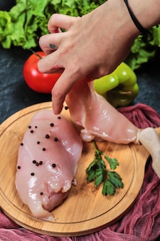 Hand holding piece of raw chicken fillet from wooden plate with fresh vegetables.