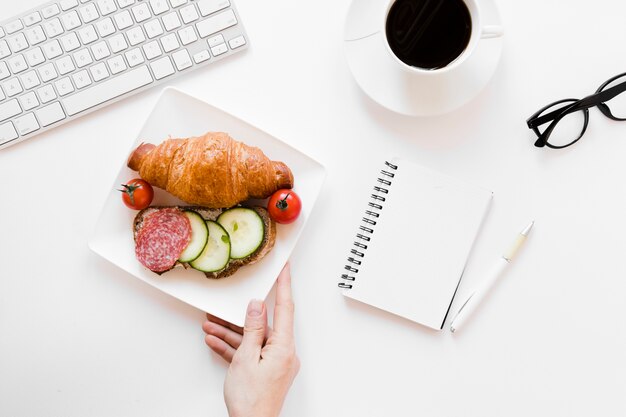 Hand holding plate with croissant and sandwich near notebook