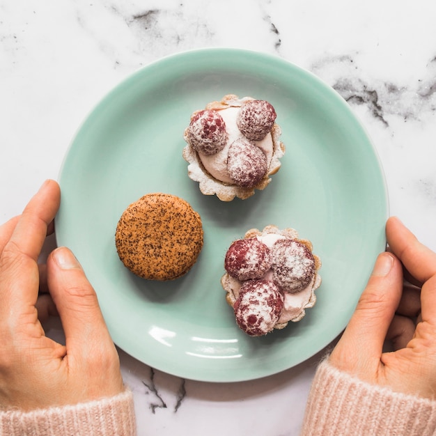 Hand holding plate with macaroon and two cup cakes on marble backdrop