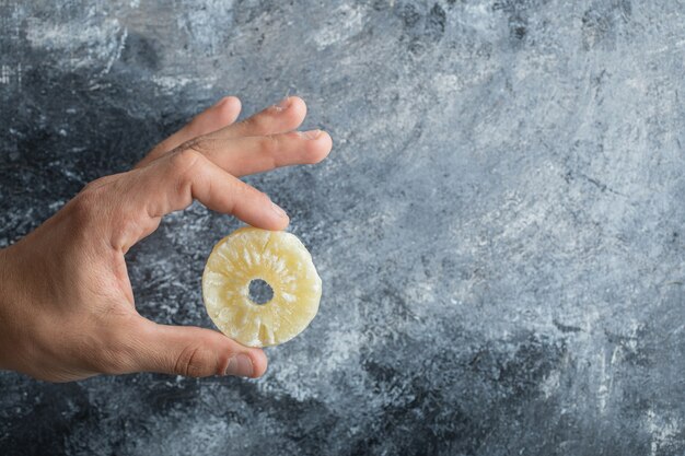 Hand holding a slice of dried pineapple on a gray background. 