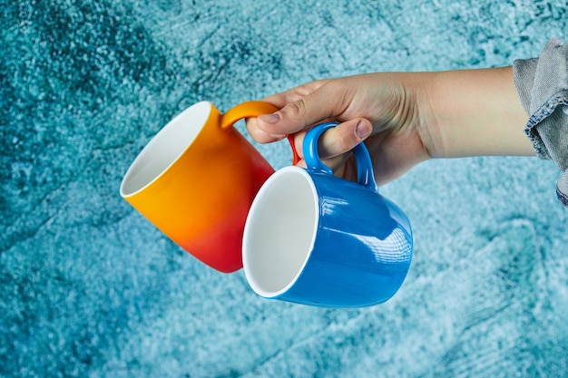 Free photo hand holding two mugs on blue background.