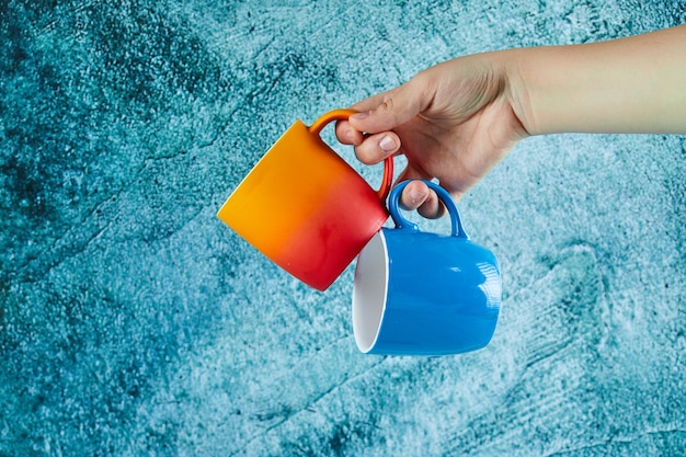 Hand holding two mugs on blue background. 