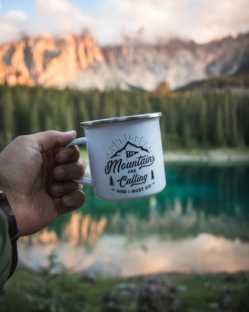 Foto gratuita mano che tiene una tazza bianca con un paesaggio di montagna sfocato come