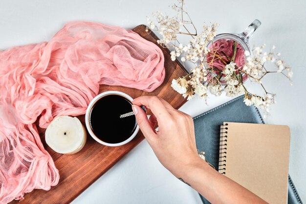 Рука держит чашку кофе на деревянной доске со свечой, тканью понк и цветами.