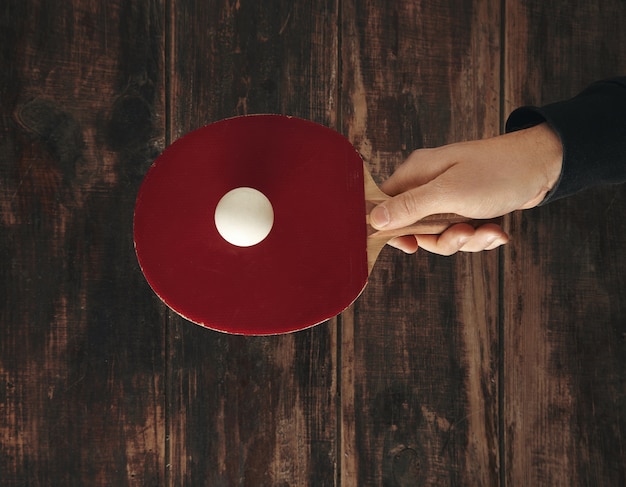 Hand holds one professional rocket above wooden aged table with ball on it and ready to play ping pong